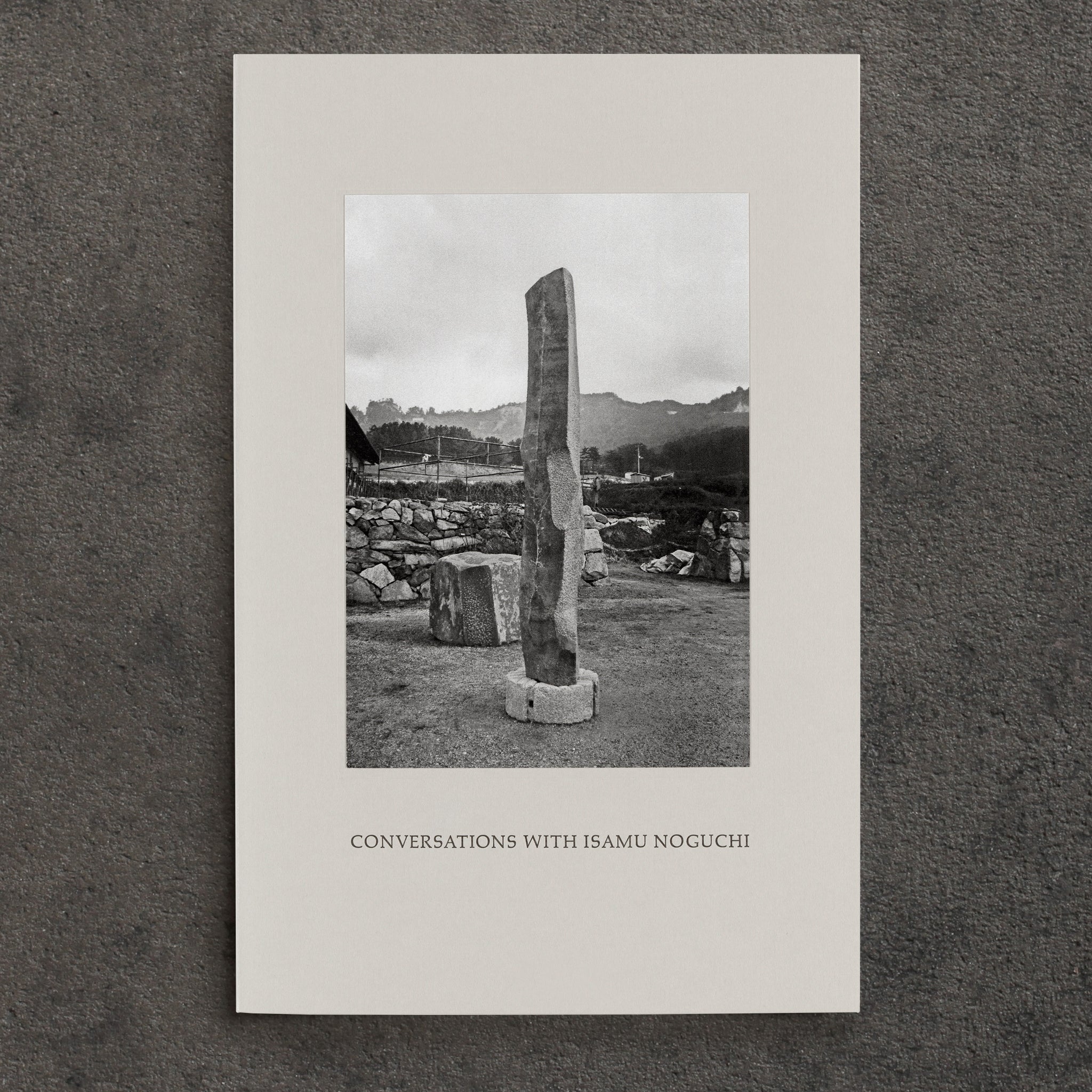 Conversations with Isamu Noguchi, front cover. Grey book cover with black and white photograph of view of Isamu Noguchi’s To Tallness and sculptures in progress at his studio in Mure, surrounded by mountains. The title "Conversations with Isamu Noguchi" is in Palatino Caps below the image, printed letterpress. 