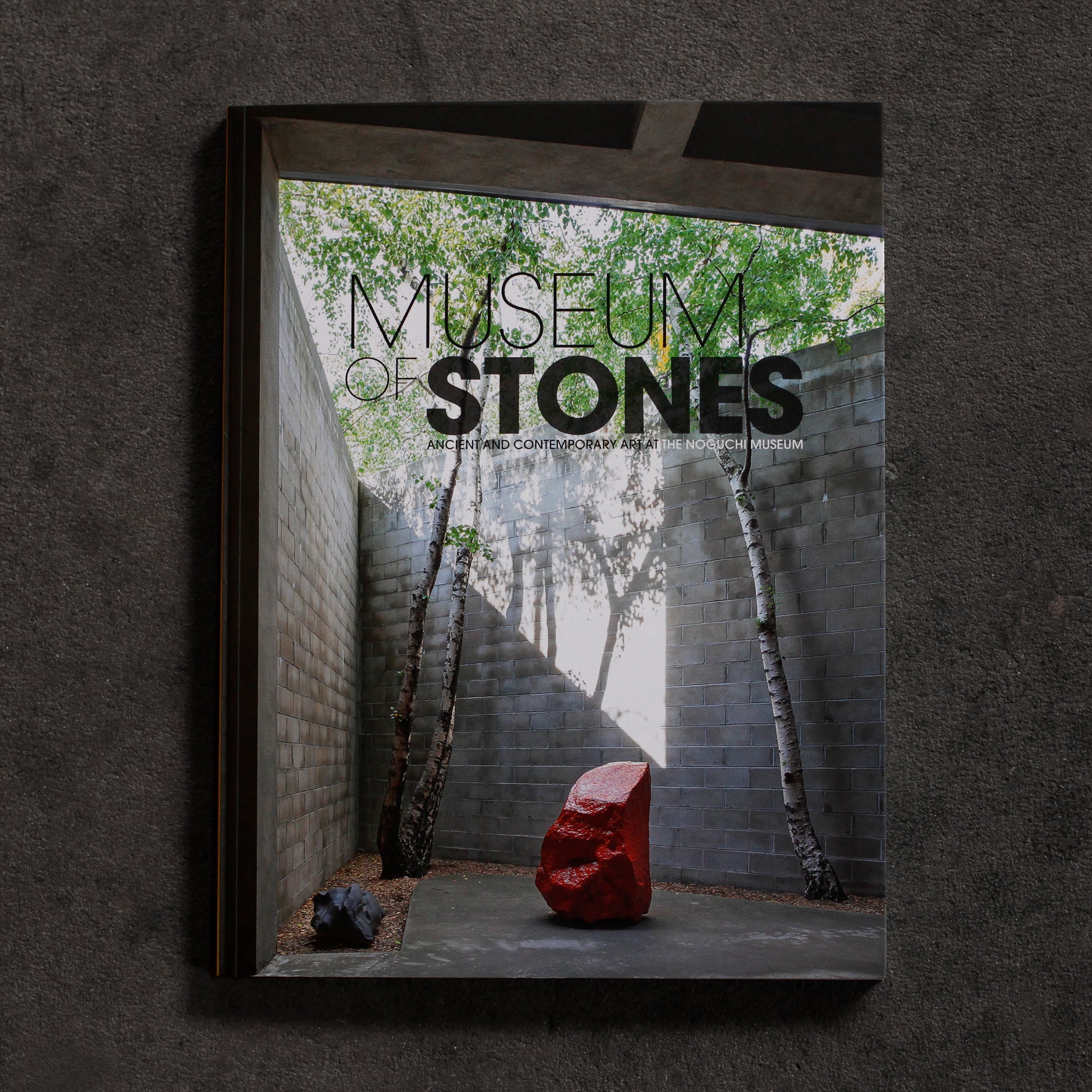 Museum of Stones cover. Bright red stone sculpture by Bosco Sodi stands between two birch trees in a beam of sunlight in the cutaway of the Noguchi Museum’s Area 1