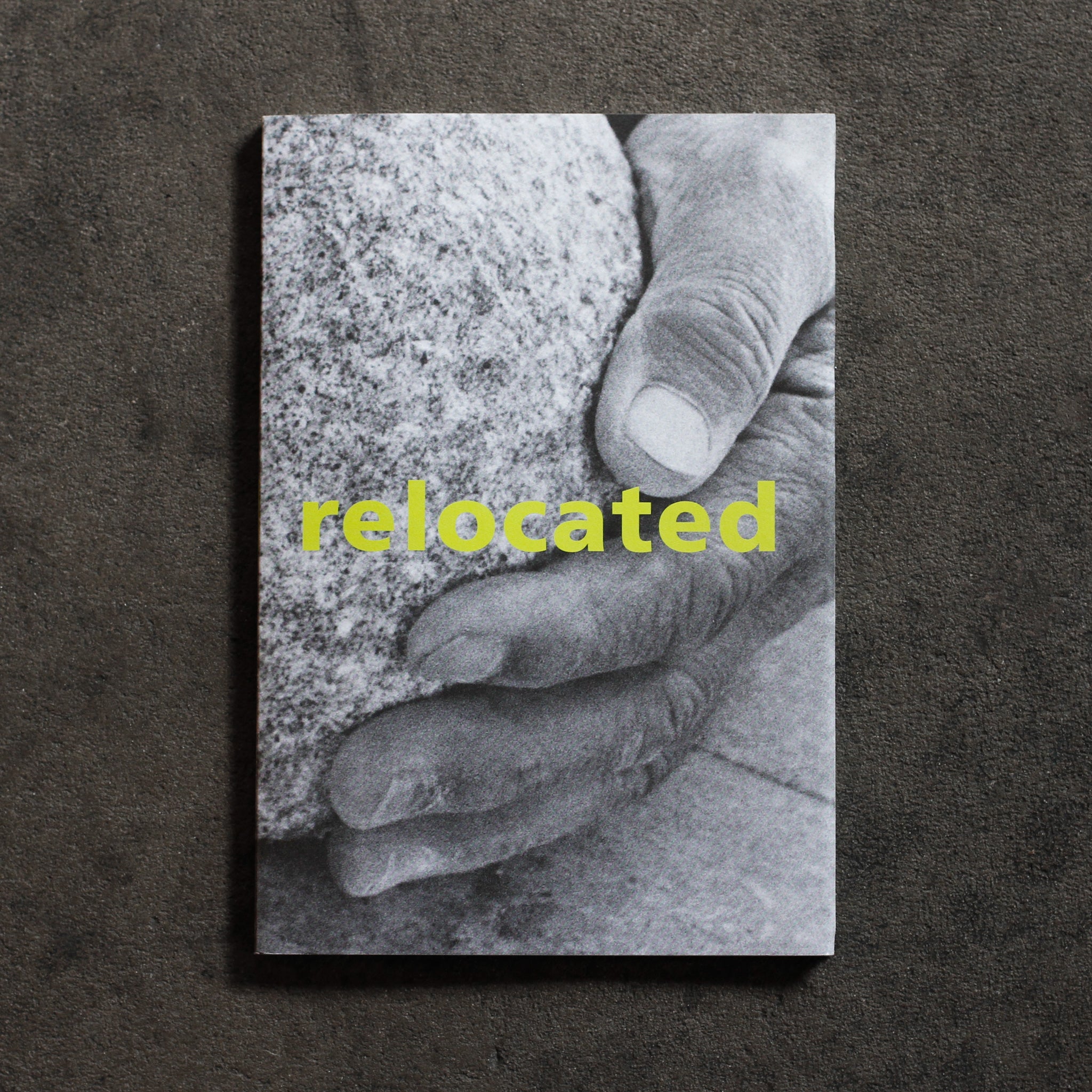 Cover of 'Relocated.' A black and white closeup of Isamu Noguchi's hand holding a chiseled stone, with large headline 'relocated' across the center of the book in large neon yellow sans serif type
