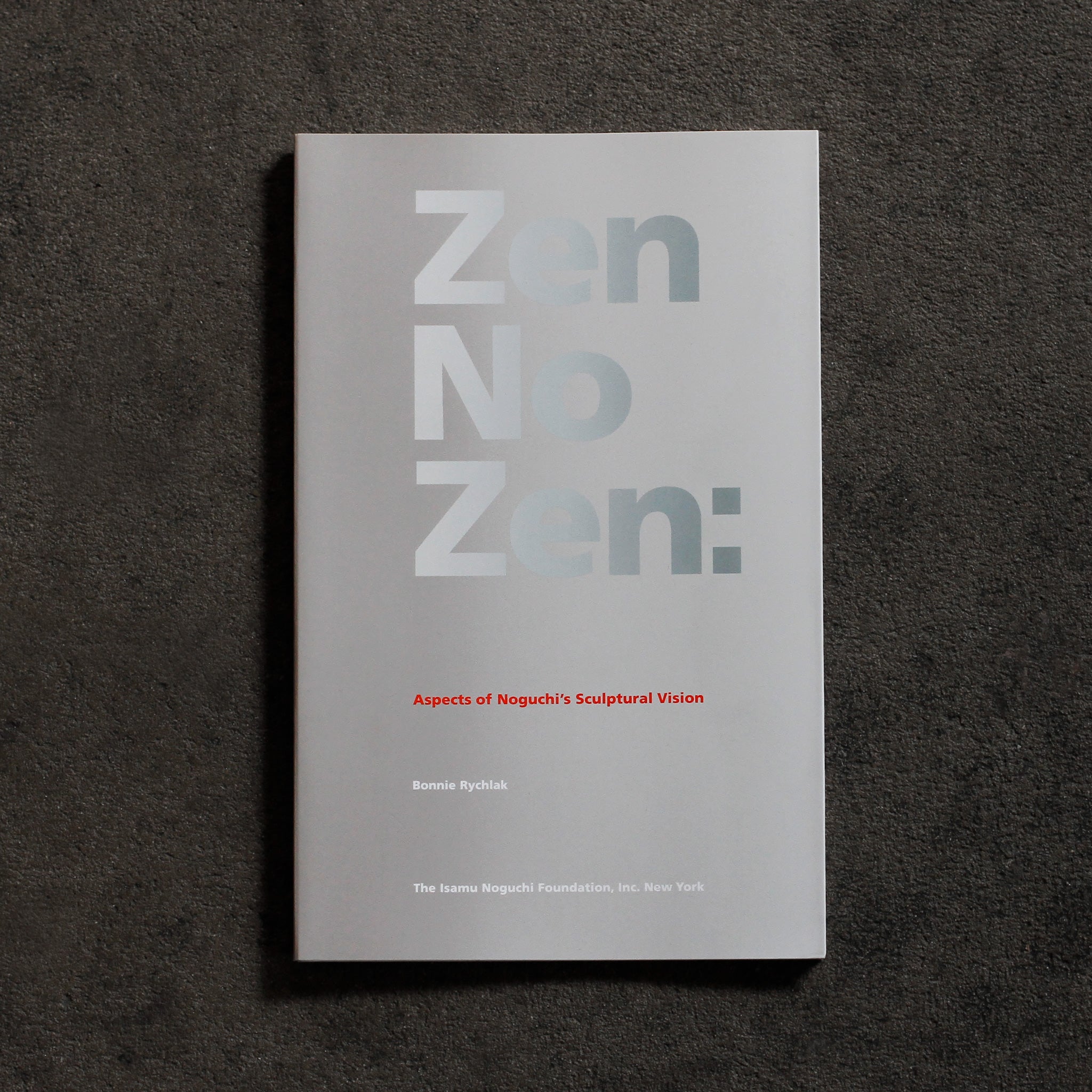 Cover of 'Zen No Zen.' Softcover book with silver grey background and large reflective silver bold sans serif headline text 'Zen No Zen'. In small red sans serif text below, 'Aspects of Noguchi’s Sculptural Vision' and in white text below, same type style, 'Bonnie Rychlak,' 'The Isamu Noguchi Foundation Inc. New York'